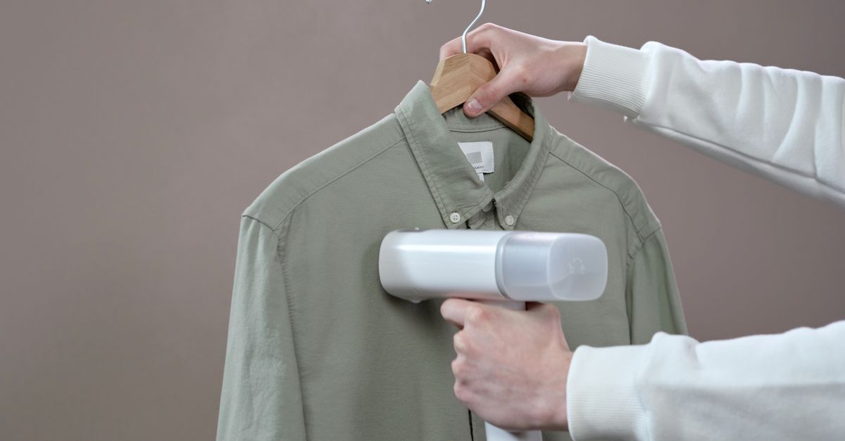 Hands steaming a green shirt with a portable steamer against a grey background.